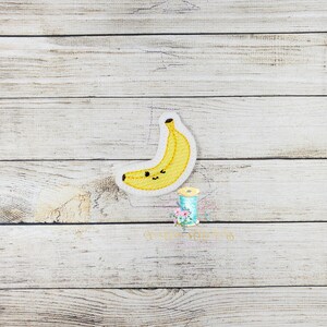 Peut inclure: Une banane en feutre jaune avec un visage souriant mignon. La banane est sur un fond en bois clair.