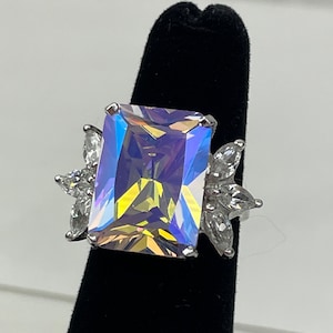 Può includere: Un anello d'argento con una grande pietra preziosa rettangolare iridescente circondata da pietre preziose più piccole e trasparenti.
