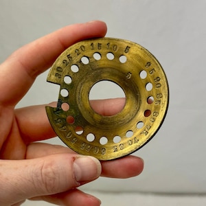 Puede incluir: Un calibre de latón con forma circular y un agujero en el centro. El calibre tiene números del 5 al 30 impresos alrededor del borde.