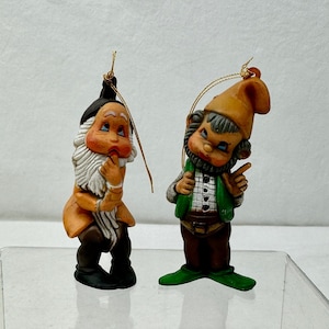 May include: Two vintage Christmas ornaments depicting gnomes. The gnome on the left is wearing a brown hat, a brown and orange jacket, and a white beard. The gnome on the right is wearing a brown hat, a green and brown jacket, and a white beard. Both gnomes are holding a gold string.