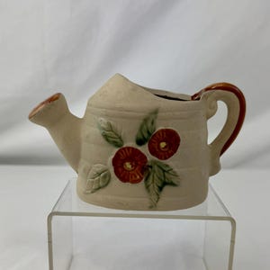 May include: A small, cream ceramic watering can with a red and green floral design. The handle and spout have brown accents. The watering can is displayed on a clear acrylic stand.