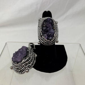 May include: Two silver rings each featuring a large purple amethyst stone set within a border of smaller clear stones.