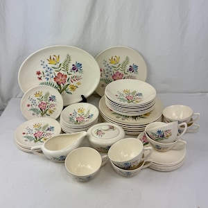 Puede incluir: Un juego de vajilla floral vintage de color crema. El juego incluye platos, platillos, tazas, azucarero, cremera y fuentes. El diseño floral presenta flores rosas, amarillas, azules y moradas con hojas verdes.