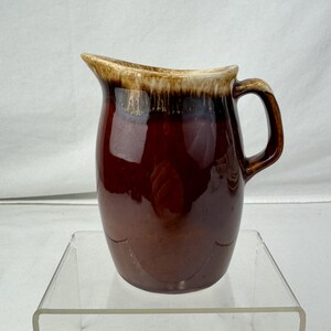 May include: A brown ceramic pitcher with a handle and a spout. The pitcher has a glossy finish and a lighter brown rim. The body of the pitcher has a curved design near the bottom. The pitcher is sitting on a clear acrylic stand.