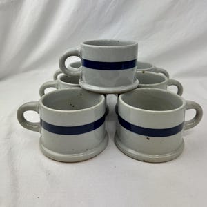 May include: A set of six small, gray ceramic mugs with a dark blue horizontal stripe. Each mug has a rounded handle and a sturdy base. The mugs are stacked in a pyramid shape, showcasing their design and form.