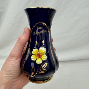 Könnte beinhalten: Eine dunkelblaue Keramikvase mit goldenen Verzierungen und Akzenten. Die Vase zeigt eine erhabene, dreidimensionale gelb-weiße Blume mit roter Mitte. Die Worte "Burg Hohenzollern" sind in goldener Schrift auf der Vase geschrieben.