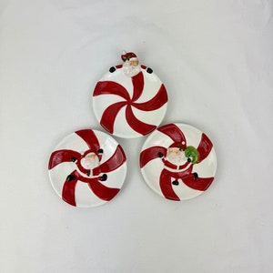 Könnte beinhalten: Drei weiße Keramik-Weihnachtsteller in Form von Zuckerstangen mit roten Streifen und einer Weihnachtsmannfigur auf jedem.