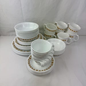 Puede incluir: Un juego de vajilla vintage blanco con un diseño floral dorado. El juego incluye cuencos, platos, platillos y tazas. Los platos están apilados en montones ordenados, mostrando la colección completa.