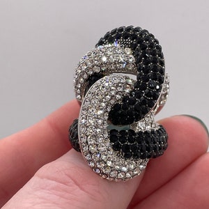 Op de afbeelding: Een zilveren ring met een zwart-wit kristalontwerp. De ring is gemaakt van twee in elkaar grijpende cirkels, een bedekt met zwarte kristallen en de andere met witte kristallen.