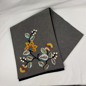 May include: A dark gray scarf with a floral embroidery design. The embroidery features yellow, orange, and blue flowers with green and teal leaves. The scarf has a black border.