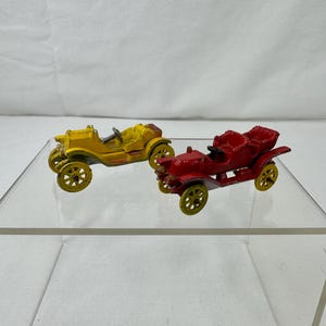 May include: Two vintage toy cars. One is yellow, the other is red. Both have yellow wheels and black seats. The cars are displayed on a clear, angled surface against a white background. The cars appear to be made of metal.