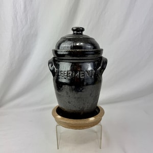 Peut inclure: Un pot de fermentation en céramique noire avec un couvercle et des poignées. Le mot "FERMENT" est en relief sur le devant. Il repose sur une base en céramique beige, exposée sur un présentoir en acrylique transparent. Le pot a une finition brillante.