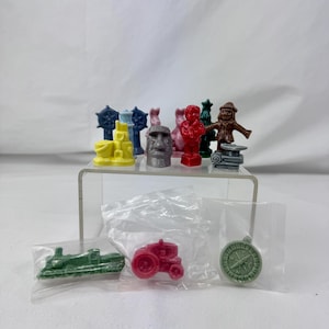 May include: A collection of small, molded game pieces in various colors and shapes, including a yellow sandcastle, a gray Moai statue, a red figure, a brown figure, a blue ship wheel, and a green tree. Three pieces are in plastic bags: a green train, a red tractor, and a light green compass.