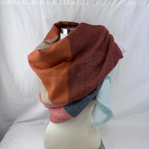 May include: A color-blocked scarf featuring sections of burnt orange, deep maroon, beige, charcoal gray, and light blue. The scarf has a soft, knit texture and includes a fringed edge on the light blue section, draped over a display form.