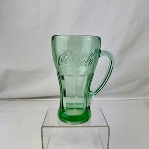 Könnte beinhalten: Ein grüner Coca-Cola-Becher aus Glas mit Henkel. Das Coca-Cola-Logo ist auf der Vorderseite eingeprägt. Der Becher steht auf einem transparenten Acrylständer vor weißem Hintergrund.