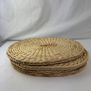 Puede incluir: Una pila de cuatro manteles individuales redondos y tejidos. Los manteles individuales están hechos de un material de fibra natural de color beige claro. Los bordes están cuidadosamente acabados y el patrón de tejido es visible. Estos manteles individuales son adecuados para el comedor y la decoración del hogar.