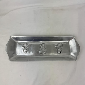 Könnte beinhalten: Ein rechteckiges, silberfarbenes Metalltablett mit drei geprägten Bienenmotiven. Das Tablett hat leicht gebogene Kanten und eine glatte Oberfläche. Die Bienenmotive sind entlang der Länge des Tabletts zentriert.