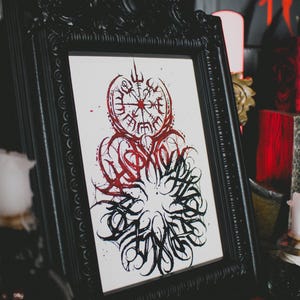 May include: A black framed print with a red and black ink illustration of a compass with a stylized design around it. The compass has a red center and black lines radiating out from the center. The stylized design around the compass is black and red and has a swirling, organic look.