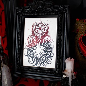 May include: A black framed print with a white background featuring a red and black ink illustration of a compass with a stylized design. The compass is surrounded by a circle of stylized lettering.