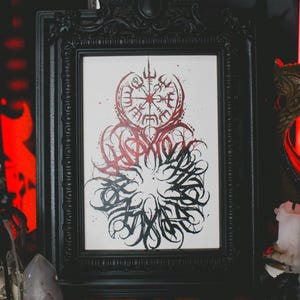 May include: A black framed print with a white background and a black and red design. The design features a circular compass with a runic symbol in the center and a surrounding circle of stylized lettering.