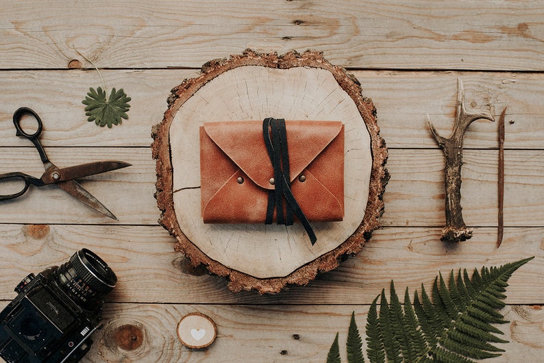 May include: A brown leather journal with a black leather strap and a wooden heart on a wooden background. The journal is surrounded by other items, including a pair of scissors, a fern, and a deer antler.