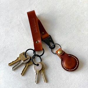 May include: A brown leather keyring with a black clasp and several keys attached. The keyring has the words "STORY IS HERE" embossed on the strap. The keyring also has a small, round leather tag.