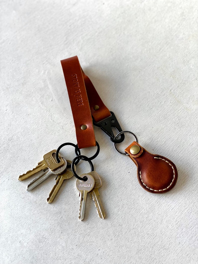 May include: A brown leather keyring with a black clasp and several keys attached. The keyring has the words "STORY IS HERE" embossed on the strap. The keyring also has a small, round leather tag.