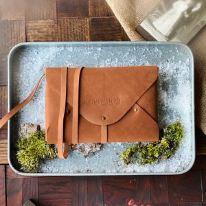 May include: A brown leather envelope with a strap closure and the text "MIZMASY STUDIO" embossed on the front.