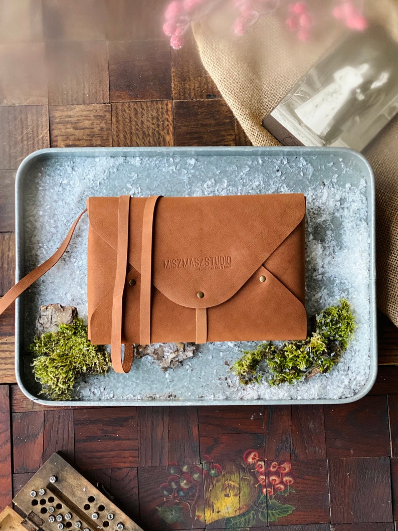May include: A brown leather envelope with a strap closure and the text "MIZMASY STUDIO" embossed on the front.