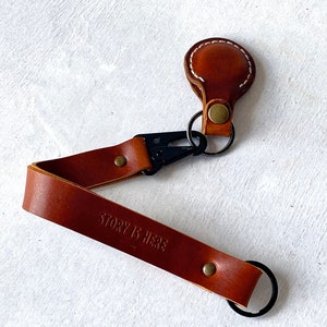 May include: A brown leather keychain with the embossed text "STORY IS HERE". The keychain has a black clasp and a small, round leather piece with white stitching. A metal ring is attached for keys.