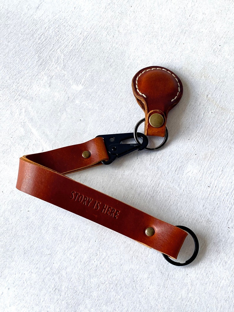May include: A brown leather keychain with the embossed text "STORY IS HERE". The keychain has a black clasp and a small, round leather piece with white stitching. A metal ring is attached for keys.