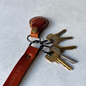 May include: A brown leather keyring with the text "STORY IS HERE" embossed on the strap. The keyring holds several metal keys. The leather is stitched with white thread and has a brass rivet.