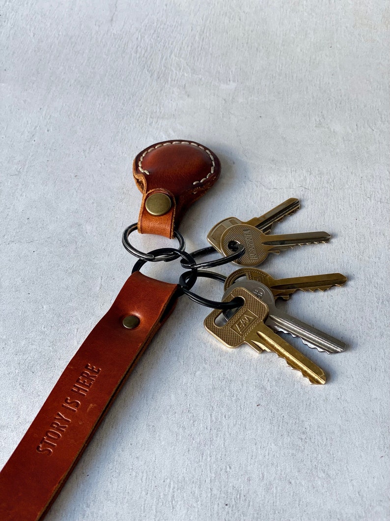 May include: A brown leather keyring with the text "STORY IS HERE" embossed on the strap. The keyring holds several metal keys. The leather is stitched with white thread and has a brass rivet.