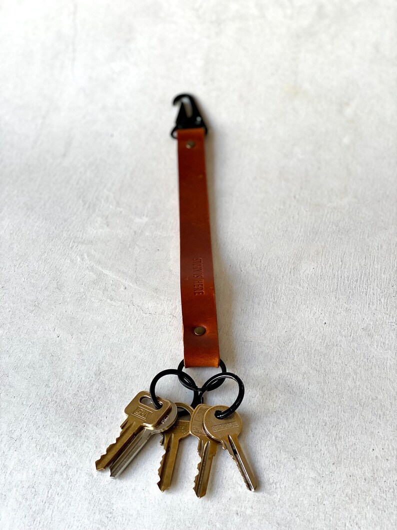 May include: A brown leather key fob with a black metal clasp and key rings, holding three keys. The text "STORY IS HERE" is stamped on the leather. The key fob is designed to keep keys organised and accessible.