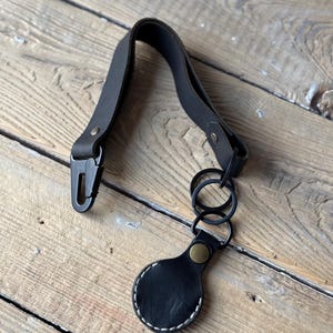 May include: Black leather key fob with a metal clasp and key rings. The fob has a circular tag with white stitching and a brass-coloured rivet. The leather strap is attached to a metal clasp.