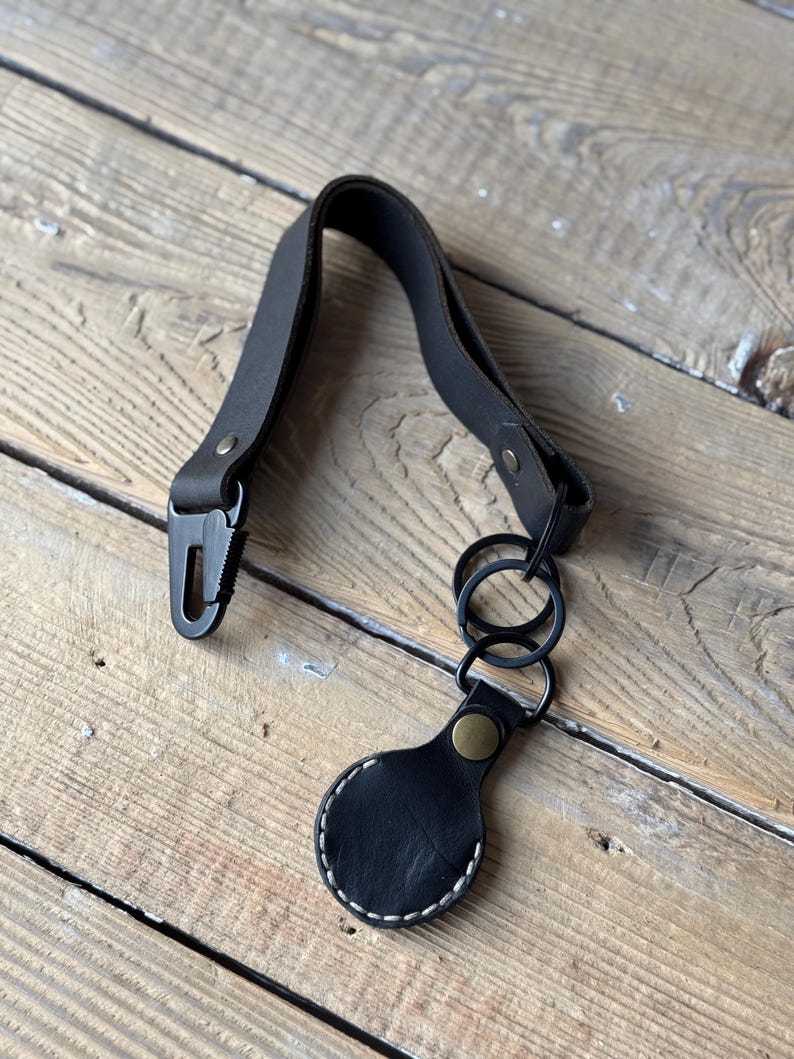 May include: Black leather key fob with a metal clasp and key rings. The fob has a circular tag with white stitching and a brass-coloured rivet. The leather strap is attached to a metal clasp.