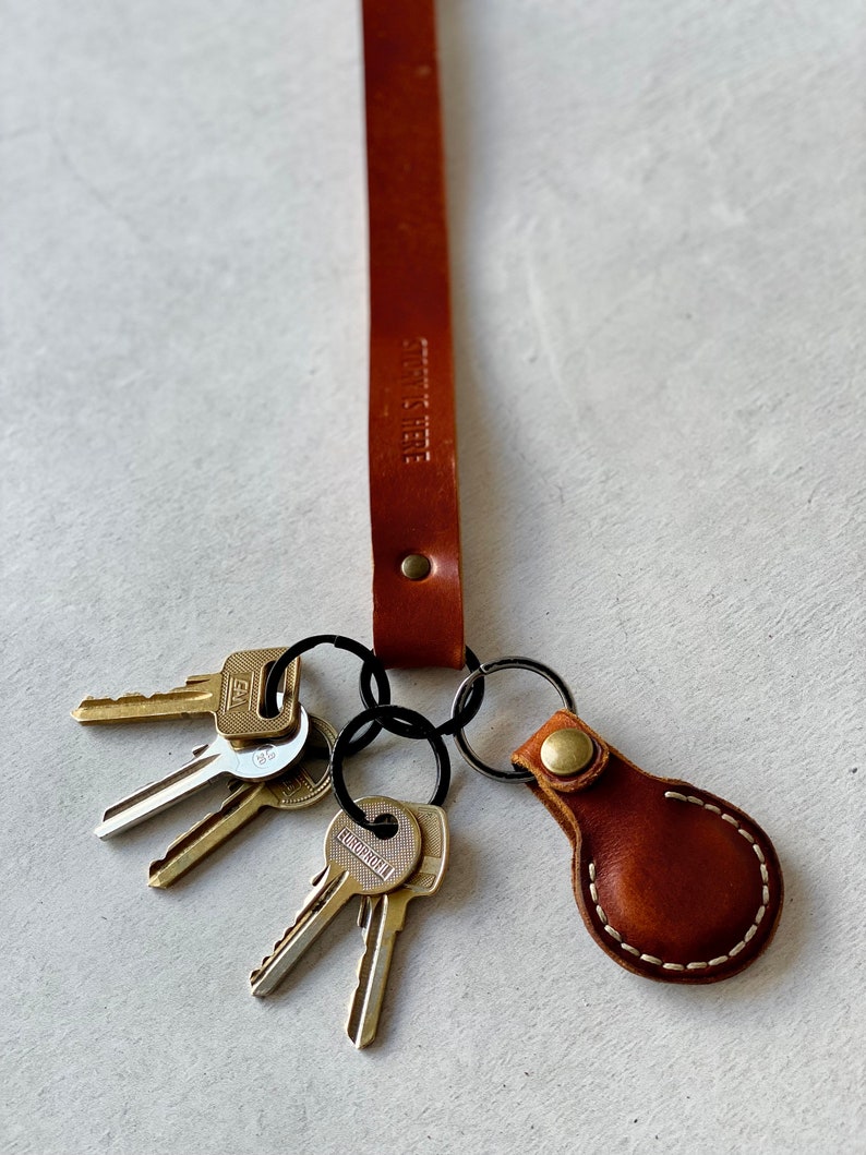 May include: A brown leather keychain with the text "STORY IS HERE" embossed on the strap. The keychain holds several keys and a small, stitched leather fob with a brass button.