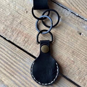 May include: Black leather keychain with a stitched border and a brass-coloured snap. The keychain has three black metal rings and a black leather strap with a brass-coloured rivet.