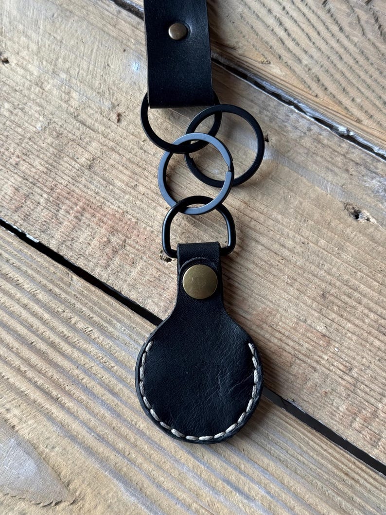 May include: Black leather keychain with a stitched border and a brass-coloured snap. The keychain has three black metal rings and a black leather strap with a brass-coloured rivet.