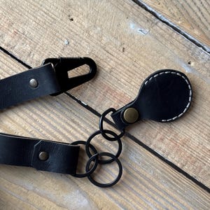 May include: A close-up shot of a black leather strap with a metal clasp and rivets. The strap is attached to black metal rings and a small, rounded leather piece with white stitching. The background is a wooden surface.