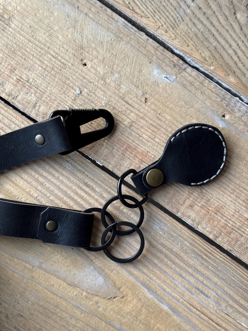 May include: A close-up shot of a black leather strap with a metal clasp and rivets. The strap is attached to black metal rings and a small, rounded leather piece with white stitching. The background is a wooden surface.