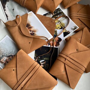 May include: A group of brown leather envelopes with a flap closure and a strap. The envelopes are arranged on a white surface with printed photographs underneath.