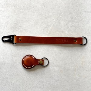 May include: Two leather keychains. One is a long strap with a black clasp and the text "STORY IS HERE". The other is a small, round keychain with white stitching.