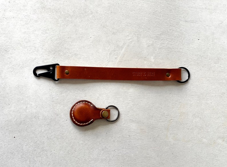May include: Two leather keychains. One is a long strap with a black clasp and the text "STORY IS HERE". The other is a small, round keychain with white stitching.