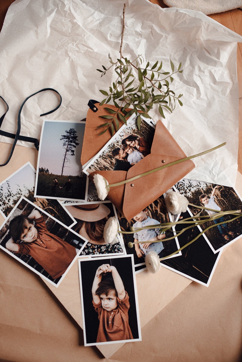 May include: A brown leather envelope with a flap and a metal clasp, filled with printed photographs of children and families. The envelope is surrounded by white flowers and green leaves.