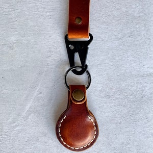 May include: A brown leather keychain with a black metal clasp and a key ring. The keychain features a stitched, oval-shaped leather tag. The leather is a warm brown colour, and the stitching is white. The hardware is black and brass-coloured.
