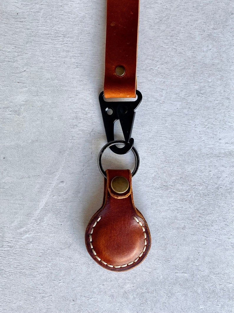 May include: A brown leather keychain with a black metal clasp and a key ring. The keychain features a stitched, oval-shaped leather tag. The leather is a warm brown colour, and the stitching is white. The hardware is black and brass-coloured.
