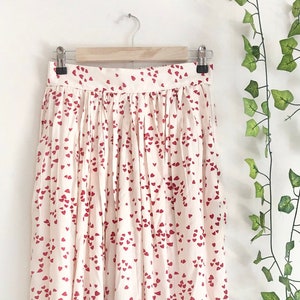 May include: A white maxi skirt with a red heart print. The skirt has a pleated design and is hanging on a hanger.