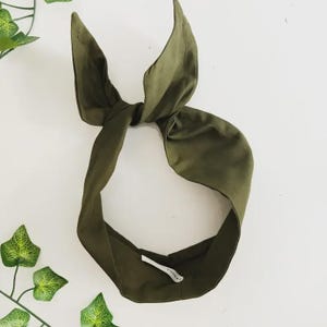 Puede incluir: Una diadema de tela color verde oliva con un diseño de nudo en la parte superior. La diadema está confeccionada con un material liso de color sólido e incluye una pequeña etiqueta blanca en el interior. Tiene forma de bucle circular.