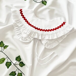 May include: White ruffled collar with a red scalloped trim. The collar is made of a soft, lightweight fabric and has a delicate, feminine look.